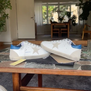 Golden Goose Superstar sneakers vit/blå Storlek 46 - Golden Goose Superstar sneakers i vitt skinn med grå snörning och beige stjärndetalj på sidan. Hälpartiet är i metallicblått och sulan är platt med svart mönstrad undersida. Klassisk rund tå och cool kontrast mellan färgerna. Kommer med dustbag och originaltag. Kommer utan skokartong Storlek 46 men sitter som 45