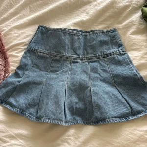 Blå jeanskjol med veckad design - Säljer en snygg blå jeanskjol med veckad nederdel och hög midja. Kjolen har klassisk denimlook och är perfekt för dig som gillar en trendig och avslappnad stil. Passar till sneakers eller boots och är lätt att matcha med olika toppar.