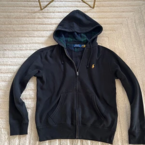 Svart hoodie från Polo Ralph Lauren - Svart hoodie från Polo Ralph Lauren med gul broderad logga på bröstet. Tröjan har huva med snörning, dragkedja framtill och två stora fickor. Insidan av huvan har ett rutigt mönster i grönt och blått. Perfekt för chill dagar.