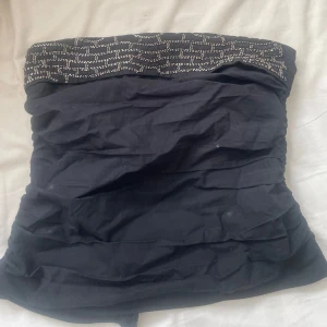 Svart tubtopp med pärldetaljer - Svart tubtopp med glittrande pärldetaljer upptill och snygg dragkedja baktill. Toppen har ett rynkat tyg som ger en cool struktur och är axelbandslös. Perfekt för att sticka ut med lite extra bling. Passar dig som gillar att mixa stil med edge.