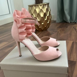 Baby Rosa pumps med volang från Bianco - Snygga rosa pumps från Bianco med hög klack och öppen tå. Skorna har en unik volangdetalj runt hälen och ett smalt ankelband med spänne. Materialet är mjuk mocka och färgen är ljusrosa, perfekt för dig som gillar att sticka ut med detaljer.