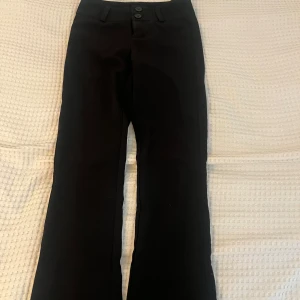Svarta bootcut kostymbyxor Gina Tricot XS - Svarta kostymbyxor från Gina Tricot i storlek XS. Snygg bootcut-modell med två knappar fram, bälteshällor och fickor med lock och knapp bak. Perfekt för dig som gillar klassisk stil med en modern twist.