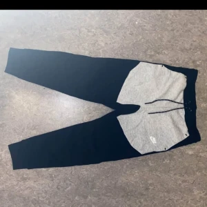 Nike mjukisbyxor svart/grå  - Nike mjukisbyxor i svart med grå panel framtill och vit logga. Byxorna har snörning i midjan, ribbade muddar och en dragkedjeficka bak. Skön passform och stilren design, perfekta för chill eller träning.