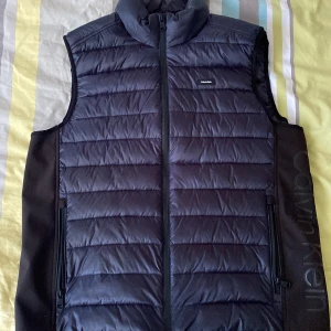 Svart dunväst från Calvin Klein XL - Snygg svart dunväst från Calvin Klein i storlek XL. Västen har quiltad framsida, hög krage och dragkedja hela vägen. Stora loggan på sidan och två praktiska fickor med dragkedja. Köptes för 1500 och kan tänka mig att gå ner lite i pris 
