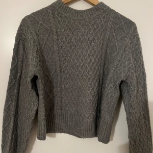Grå stickad tröja från H&M - Säljer en grå stickad tröja från H&M i storlek XS. Tröjan har ett snyggt kabelstickat mönster med ribbad kant vid hals, ärmslut och nederkant. Perfekt för dig som gillar klassisk stil med lite edge. Långärmad och riktigt skön och varm att ha på sig under kyliga dagar. 