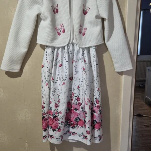 Vit långklänning med blommor och fjärilar - Superfin vit långklänning med rosa och röda blommor samt fjärilar på kjolen och överdelen. Kjolen har ett romantiskt blommönster och överdelen är dekorerad med broderade fjärilar. Klänningen har långärmad överdel och är gjord i mjukt, lätt material. Perfekt för vår och sommar!