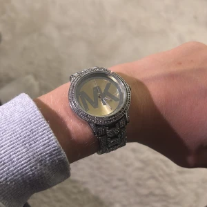 Michael Kors glittrig silverklocka - Säljer en snygg klocka från Michael Kors i silver med glittriga stenar runt urtavlan och på länken. Stor MK-logga på den guldtonade urtavlan. Länken har coola detaljer och ett rejält spänne. Perfekt för dig som gillar bling och vill sticka ut.