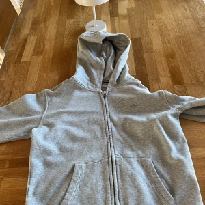 Grå hoodie med dragkedja och huva - Säljer en chill grå hoodie från Eton med dragkedja framtill och stor huva. Tröjan har två rejäla fickor och ett litet broderat Eton-emblem på bröstet. Perfekt för att slappa eller dra på sig när det är lite kyligt ute.            I PRINCIP NYANVÄND