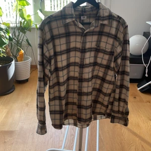Beige rutig skjorta från Uniqlo - Snygg beige skjorta från Uniqlo med rutigt mönster i mörkbrunt och blått. Skjortan har klassisk krage, bröstficka och knappar framtill. Perfekt för dig som gillar en chill och avslappnad stil. Materialet är mjuk bomull och modellen är långärmad. Storlek XS men sitter mer som en S, skriv vid funderingar 