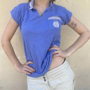 Blå pikétröja från Lacoste med mönster - Blå pikétröja från Lacoste med korta ärmar och klassisk krage med ränder i vitt och beige. Bröstfickan har ett snyggt argylemönster i vitt och beige, och den ikoniska krokodilloggan sitter ovanför fickan. Perfekt för dig som gillar sportig och retro stil.