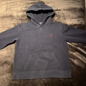 Mörkblå hoodie från Polo Ralph Lauren - Snygg mörkblå hoodie från Polo Ralph Lauren med klassisk röd broderad logga på bröstet. Tröjan har huva, känguruficka och långa ärmar. Perfekt för chill dagar och enkel att matcha med jeans eller joggers. Bra kvalitet.