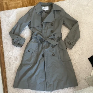 Grå trenchcoat Designers Remix stl 36 - Snygg grå trenchcoat från Designers Remix Collection i storlek 36. Jackan har klassisk dubbelknäppning, stora fickor och bälte i midjan. Långa ärmar med knappar vid mudden och stilren krage. Perfekt för dig som gillar minimalistisk och cool stil.