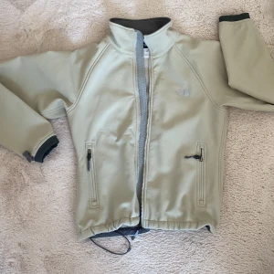 Beige vindjacka från The North Face - Snygg beige vindjacka från The North Face i storlek M. Jackan har hög krage, två fickor med dragkedja och justerbar nederkant med snörning. Materialet är nylon och elastan, vilket gör den både lätt och smidig. Perfekt för dig som gillar stilren och funktionell design.
