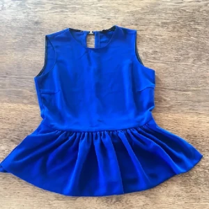 Zara Kornblå ärmlös blus med volang - Supersnygg kornblå blus med volangkant nedtill och rund hals. Blusen är ärmlös och har en rak passform med knäppning i ryggen. Materialet känns mjukt och har ett lätt glansigt utseende, perfekt för att sticka ut. Volangen ger en extra touch av stil.