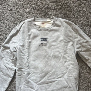 Grå sweatshirt från Levi's med tryck - Säljer en grå sweatshirt från Levi's i storlek S. Tröjan har ett stort Levi's-tryck på ryggen och mindre tryck framtill. Rund hals och långa ärmar. Mjuk bomullskvalitet som är skön att ha på sig. 