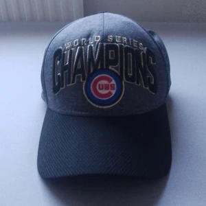 Chicago Cubs World Series Champions keps - Snygg keps från New Era med Chicago Cubs World Series Champions broderat i guld och svart. Framsidan har Cubs-loggan i rött, blått och vitt. Kepsen är mörkgrå med svart skärm och har broderade detaljer på sidorna. Perfekt för dig som älskar baseball och vill sticka ut.