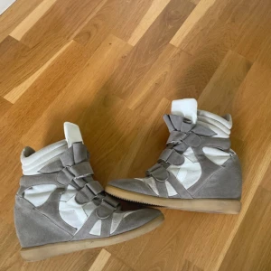 ISABEL MARANT LIKNANDE - Säljer ett par grå och vita wedge sneakers med högt skaft och coola kardborreband. Skorna har en plattformssula och är tillverkade i mocka och syntetmaterial. De har en rund tå och en chunky look som verkligen sticker ut. Perfekta för dig som gillar streetstyle och vill ha extra höjd.