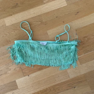 Mintgrön fransig bandeau topp - Supercool mintgrön bandeau topp från na-kd/hanna schönberg med tunna axelband och massor av fransar som ger en unik och lekfull vibe. Toppen är croppad och har en mjuk syntetisk känsla, perfekt för att sticka ut med färg och rörelse.