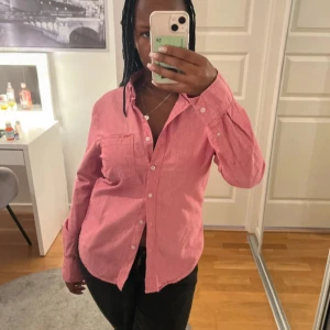 Rosa långärmad skjorta med knappar - Säljer en stilren rosa skjorta med vita knappar framtill. Skjortan har en bröstficka och lång ärm, passformen är normal och materialet känns som bomull, perfekt för en chill och snygg look.
