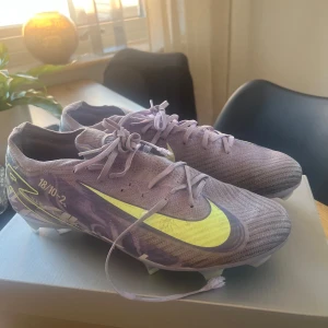 Lila Nike fotbollsskor med neon-detaljer - Säljer ett par lila Nike fotbollsskor med neon-gula detaljer och snörning. Endast använda en månad men har legat i garderoben sen en stämpling som gjorde hål i snörningen!