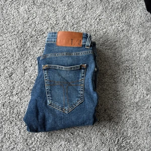 Blå jeans från tiger of Sweden 28/30 - Säljer ett par blå skinny jeans från T/J, modell Slight Spike, tillverkade i Italien. Jeansen har klassisk femficksdesign, bruna detaljer och en snygg läderpatch bak i midjan. Passformen är tight och materialet är slitstarkt denim.