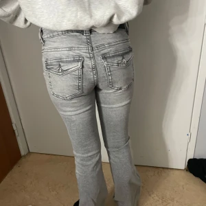Grå bootcut jeans med fickdetaljer - Snygga grå jeans med bootcut passform och fickor med lock och knapp bak. Jeansen har låg midja och är gjorda i klassiskt denimtyg. Använt 1 gång som nya💕pris kan diskuteras, köpt för 600kr