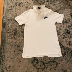 Vit Nike t-shirt med krage/ piké  - Snygg vit t-shirt från Nike med klassisk krage och korta ärmar. Svart Nike-logga på bröstet ger en sportig vibe. Perfekt för dig som gillar enkel och stilren design. Materialet känns mjukt och skönt mot huden.