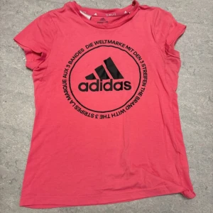 Rosa Adidas t-shirt med tryck - Snygg rosa t-shirt från Adidas med stort svart logotryck på bröstet och rund hals. Modellen är kortärmad och tillverkad i mjuk bomull. Perfekt för dig som gillar sportig stil och vill sticka ut med färg. Den är i L men passar definitivt xs/s