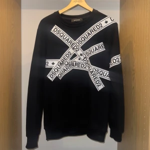 Svart sweatshirt från Dsquared2  - Svart sweatshirt från Dsquared2 i storlek L med vita tryckta band i kors över bröstet där det står DSQUARED2. Tröjan har rund halsringning och långa ärmar. Tillverkad i bomull och har en clean, modern look med tydlig logga framtill.
