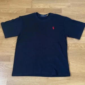 Mörkblå t-shirt från Polo Ralph Lauren - Snygg mörkblå t-shirt från Polo Ralph Lauren med klassisk röd logobrodyr på bröstet. Modellen har rund hals och korta ärmar, perfekt för en clean och stilren look. Tillverkad i mjuk bomull som känns skön mot huden.