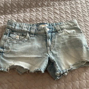 Ljusblå jeansshorts med broderade fickor - Snygga ljusblå jeansshorts från Gina Tricot med råa kanter och slitningar för en cool vibe. Baksidan har två fickor med detaljerad paisleybroderi i beige och grått. Klassisk femficksmodell, knapp och dragkedja framtill. Perfekta för sommaren! Dom är nästan helt nya har aldrig använts! Från början kostat 359 kr