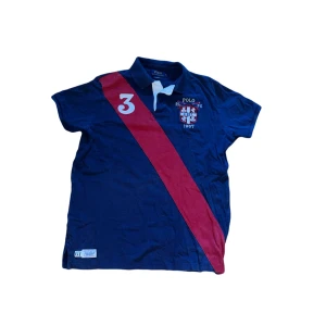 Ralph Lauren piké  - Snygg mörkblå t-shirt från Polo Ralph Lauren med klassisk krage och korta ärmar. Framsidan har en bred röd diagonal rand, vit siffra 3 och broderad logga med sköld. Materialet är bomull och passformen är normal. Perfekt för dig som gillar sportig och stilren look.  Jag skickar ej bild när jag bär plaggen och jag vet ej vilken längd och vikt de passar då jag inte prövar kläderna jag säljer, så för storlek hänvisar jag till måtten och passform att man googlar på modellnamn etc 😃