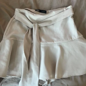 Beige kortkjol med knyt från Zara - Superfin beige kortkjol från Zara med volang och bred knyt i midjan. Kjolen har även inbyggda shorts.                Den har en liten fläck på bandet men borde gå bort i tvätten! 