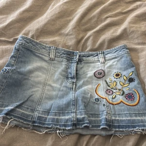 Jeanskjol med cool design - Säljer en ljusblå jeanskjol med broderade blommor i lila, gult och rosa på framsidan.  Kjolen är köpt secondhand och inte använd sen jag köpte den då den är för stor för mig. Väldigt bra skick. 