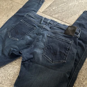 Replay Anbass jeans blå 30/34 - Snygga mörkblå Replay Anbass jeans med klassisk femficksdesign och raka ben. Jeansen har en stilren look med diskreta sömmar och läderpatch bak i midjan. Tillverkade i mjukt jeansmaterial som sitter skönt hela dagen.