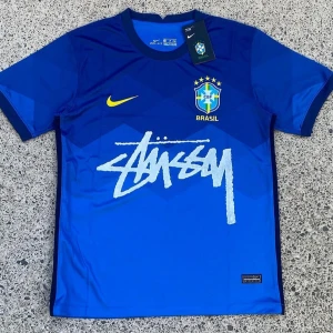 Blå Brasil Stüssy x Nike fotbollströja - Snygg blå Brasil fotbollströja från Nike i samarbete med Stüssy. Tröjan har korta ärmar, Brasil-logga med stjärnor på bröstet, gul Nike swoosh och stort Stüssy-tryck i vitt över magen. Tillverkad i lätt och ventilerande material, perfekt för match eller streetstyle.