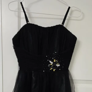 Svart kort klänning med pärldetalj - Snygg svart  klänning med tunna axelband och voluminös kjol i tyll. Livet har draperad effekt och en dekorativ pärl- och paljettblomma i midjan med svarta, vita och gula stenar. Perfekt för dig som vill sticka ut med en elegant look. Det är fest klänning som passar för nyår, bal och massa andra sorts fester som förlovnings fest