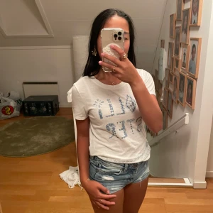 Vit Hollister t-shirt med blått tryck - Vit t-shirt från Hollister med blått tryck och mönstrad logga framtill. Materialet är mjuk bomull som känns skönt mot huden. Passformen är normal och t-shirten har ett enkelt, fräscht utseende med diskreta blå detaljer längst ner.