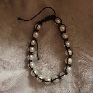 Svart pärlarmband med knyt - Snyggt handgjort armband med vita pärlor och svart snöre. Armbandet har justerbar knytning så det passar de flesta handleder. Perfekt för dig som gillar stilrena accessoarer med lite edge. Pärlorna är runda och ger en klassisk vibe till din look.