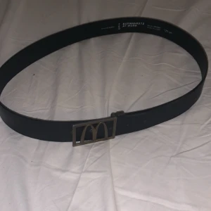 Svart skinnbälte med McDonald's-spänne - Coolt svart bälte i äkta skinn från BernhardtZ at Work med ett unikt metallspänne som har McDonald's ikoniska M-logga. Bältet är stilrent och har en rektangulär form på spännet som ger en edgy vibe. Perfekt för dig som vill sticka ut med en rolig detalj.