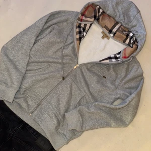 Grå Burberry hoodie med dragkedja - Säljer en grå hoodie från Burberry i storlek M. Dragkedjan är lite knepig men funkar såklart utmärkt, taggen kommer med i köpet men är avdragen. Använt hoodien 1 gång så iprincipt helt ny fortfarande.
