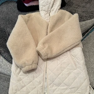 Beige teddyjacka och svart kappa - Säljer en fluffig beige teddyjacka med quiltad nederdel och huva, samt en svart kappa med bälte och snygg krage från Reera BY. Teddyjackan har dragkedja och långärmade ärmar, medan kappan har omlottstängning och är helsvart med slät yta.