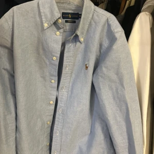 Ljusblå skjorta från Ralph Lauren - Snygg ljusblå skjorta från Ralph Lauren med klassisk button-down krage och broderad logga på bröstet. Skjortan har långa ärmar och vita knappar framtill. Perfekt för dig som gillar stilrena och tidlösa plagg med premiumkänsla.
