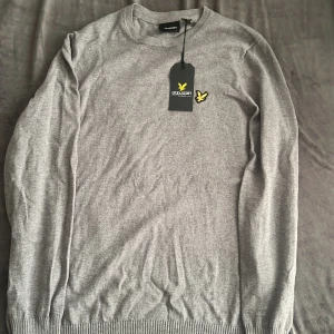 Grå stickad tröja från Lyle & Scott - Snygg grå stickad tröja från Lyle & Scott med klassisk rund hals och långärmad passform. Tröjan har en gul broderad fågel-logga på bröstet och ribbade muddar vid ärmslut och nederkant. Perfekt för dig som gillar stilrena och enkla plagg.