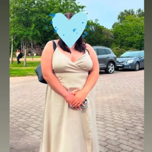 Champagnefärgad långklänning med omlott - Säljer en elegant långklänning i champagnefärg med omlottdesign och smala axelband. Klänningen har v-ringad urringning och ett mjukt, glansigt material som ger en lyxig känsla. Perfekt för dig som vill ha en stilren och feminin look.