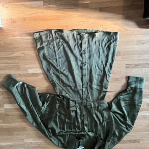 Olivgrön långklänning med knappar - Snygg olivgrön långklänning med hel knäppning framtill och långa ärmar. Klänningen har en skjortkrage och är tillverkad i ett mjukt, glansigt material som ger en cool och avslappnad vibe. Perfekt för dig som gillar stilrena och enkla plagg med lite edge.