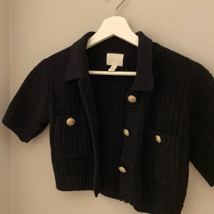 Svart stickad kofta från H&M - Säljer en svart stickad kofta från H&M med korta ärmar och dekorativa knappar i guld. Koftan har en klassisk krage och två fickor framtill. Perfekt för vardagen eller lite finare tillfällen