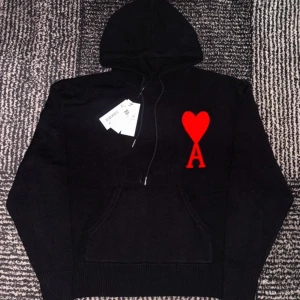 Svart hoodie med rött hjärta från Ami Paris - Säljer en stilren svart hoodie från Ami Paris med ett stort rött hjärta och bokstaven 'A' på bröstet. Tröjan har en klassisk känguruficka och en bekväm huva med dragsko. Perfekt för en avslappnad look.