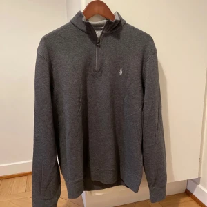 Grå tröja från Polo Ralph Lauren - Säljer en stilren grå tröja från Polo Ralph Lauren med dragkedja vid halsen. Tröjan har långa ärmar och en liten broderad logga på bröstet. Perfekt för en avslappnad och snygg look.