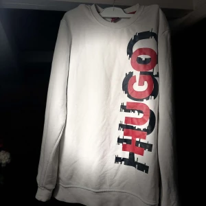 Vit långärmad tröja från Hugo Boss - Säljer en stilren vit långärmad tröja från Hugo med ett stort tryck i rött och svart på framsidan. Perfekt för en avslappnad look. Tröjan har en rund halsringning och är tillverkad i ett mjukt material.